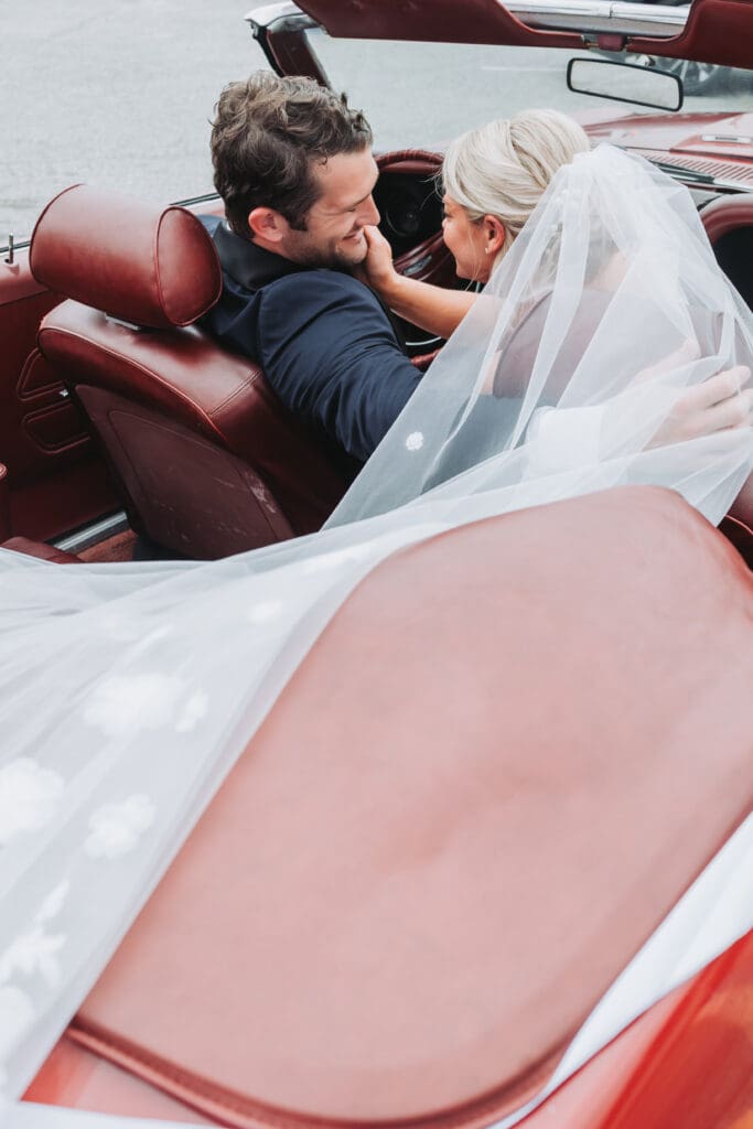 A bride and groom smiling at one another in a convertible