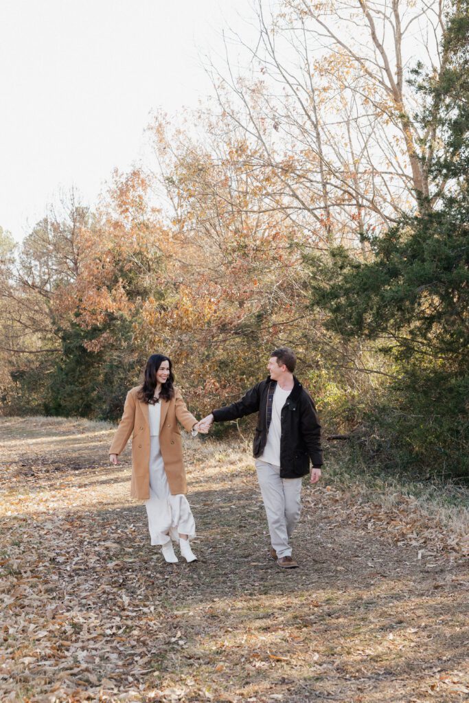Couple holding hands and smiling at each other in a grassy field at Bells Mill Park, wearing soft neutral clothing.”